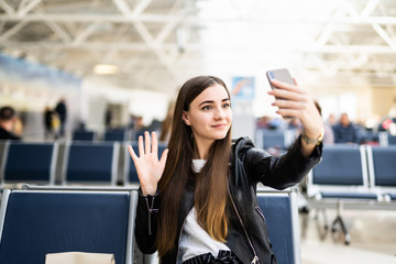 Young woman at international airport, making selfie with mobile phone and waiting for her flight. Female passenger at departure terminal, indoors.