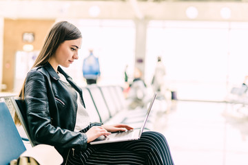 Airport Young female passenger with laptop sitting in terminal hall while waiting for her flight. Air travel concept with young casual woman sitting with hand luggage suitcase.