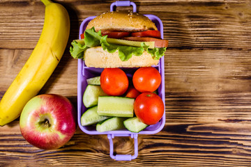Ripe apple, banana and lunch box with hamburger, cucumbers and tomatoes on wooden table. Top view