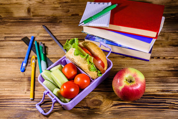 Notepad, pens, ripe apple, stack of books and lunch box with hamburger, cucumbers and tomatoes on wooden table