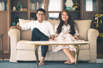Asian male and female couples, in which men wear white shirts and women wear floral dresses, smiling while sitting on a sofa in a warm home.