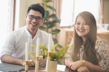 Asian male and female couples who wear white shirts and women wear floral dresses while smiling and sitting at the kitchen table.