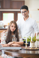 Asian male and female couples who wear white shirts and women wear floral dresses while smiling and sitting at the kitchen table.