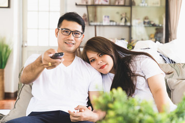 Asian male and female couples are watching TV on the sofa in the house.