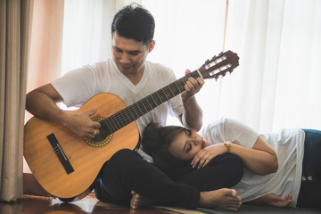 Young Asian couple wearing a white T-shirt is playing the guitar and singing together in a warm home.