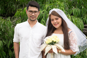 Portrait of young Asian wedding couple smiling and holding flowers in park.