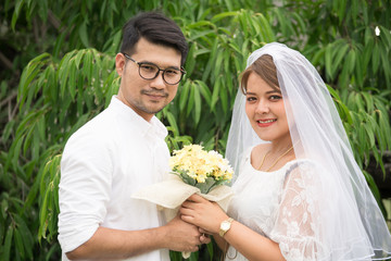 Portrait of young Asian wedding couple smiling and holding flowers in park.