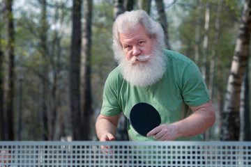 Active elderly man playing table tennis (ping pong) in park. Sunny outdoor lifestyle portrait....