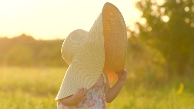 Adorable little baby girl on a field, enjoying nature outdoors wearing wide brimmed straw hat. Beautiful cute little funny one year old child smiling and doing her first steps. Slow motion 4K UHD
