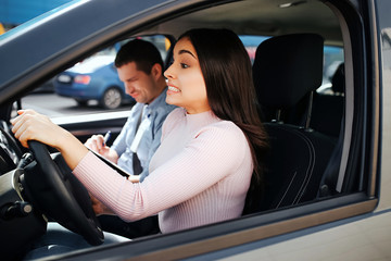 Male auto instructor takes exam in young woman. Brunette look confused straight on road and drive car. Hold hands on steering wheel. Instructor look down and write.