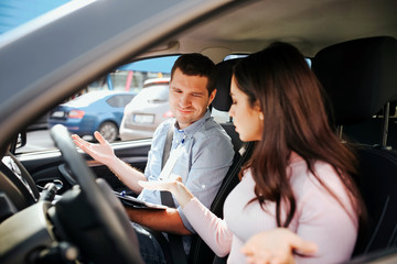 Male auto instructor takes exam in young woman. Conflict in car. People wave with hands. Look down in same direction. Sit in car.