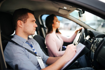 Male auto instructor takes exam in young woman. Man look forward and write. Young woman driving car careful and with attention.