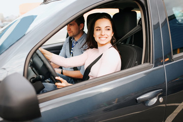 Male auto instructor takes exam in young woman. Cheerful positive and happy young woman driving car nd smile. Instructir sit besides and examine her.