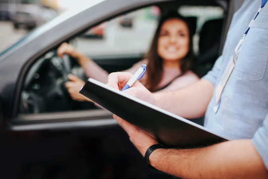 Male auto instructor takes exam in young woman. Blurred background of model sitting in car and holding hands on steering wheel. Guy hold folder with paper in hands.