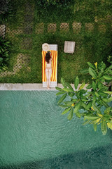 Top view of unrecognizable slim young woman in beige bikini relaxing and sunbathe near luxury swimming pool in green tropic in Ubud.Vacation concept. Drone photo