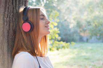 Portrait of red haired teenage girl listening to music in big pink earphones outdoors leaning to a...