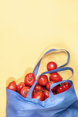 Blue non-woven shopping bag full of fresh red ripe farm tomatos and pepper on yellow background with copy space. 