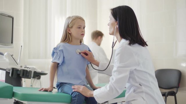Female pediatrician with stethoscope listening to lung sounds of cute little girl sitting on couch in doctors office and breathing actively, tracking medium shot