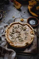 pumpkin pie decorated with pumpkin seeds on metal tray with textile, fresh pumpkin slices, seeds and fall leaves