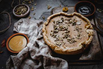 pumpkin pie decorated with pumpkin seeds on metal tray with textile, fresh pumpkin slices, seeds and fall leaves