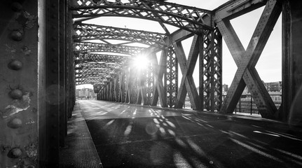 Black and white asphalt road under the steel construction of a bridge in the city on a sunny day. Evening urban scene with the sunbeam in the tunnel. City life, transport and traffic concept.