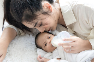Obraz na płótnie Canvas Young Asian mother hugging a cute newborn baby and feeding milk form bottle in white bedroom. Mother and child concept