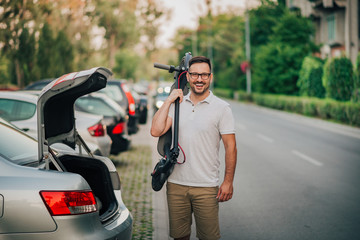 Portrait of a cheerful man standing near the car with folded electric scooter on his shoulder. © bnenin