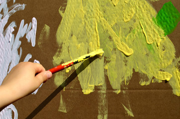 abstract yellow background with a toddler hand holding a brush and coloring a cardboard house on sunny day