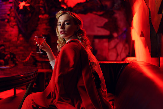 Picture of young blonde in red jacket looking at camera with cocktail in her hand in nightclub