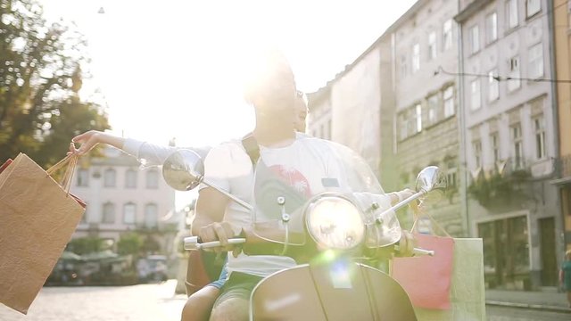Attractive young couple riding on scooter on the urban street and happy woman raised hands with purchases