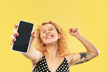 Caucasian young woman's half-length portrait on yellow studio background. Beautiful female model in black dress. Concept of human emotions, facial expression. Showing phone's screen, looks crazy happy