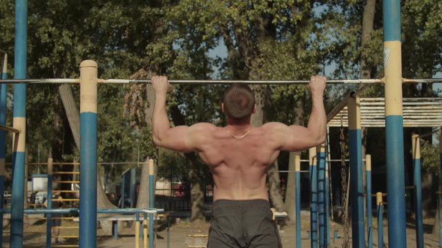 Rear view of brutal athletic fit man with perfect trained body making pull-up exercises on crossbar on sport facilities. Shirtless bodybuilder doing pull-up, strength exercise for upper body outdoors