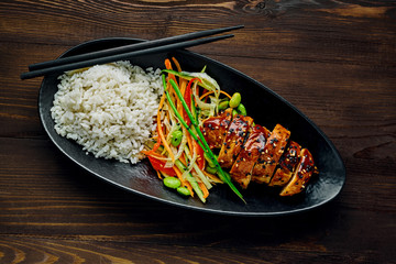 Teriyaki chicken with cucumber, ginger, cabbage and carrot salad, edamame soy beans and basmati rice in a black pottery dish. Top view, directly above.