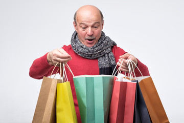 Senior hispanic man holding colored shopping bag and looking with surprise inside.