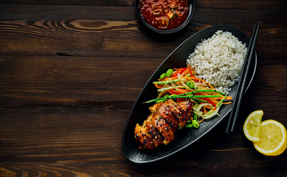 Teriyaki chicken with cucumber, ginger, cabbage and carrot salad, edamame soy beans and basmati rice in a black pottery dish with sweet chili sauce and lemon. Top view, directly above.