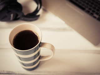 White wooden working desk with notebook, headphones and large mug of coffee. Color toned, shallow depth of field.