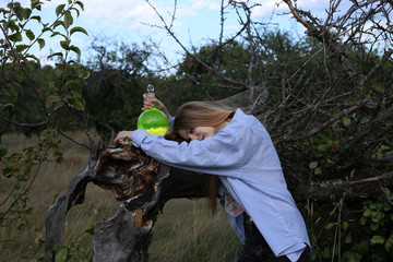 A cute girl in a man’s shirt is tired of science, holding a flask with green liquid in her hands, put it on a fallen apple tree.