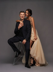 Fashion style studio portrait of young adult couple. Pretty sexy lady in luxury elegant evening dress standing near man in black suit sitting on high chair.