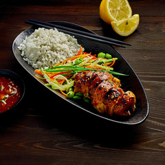 Asian- style chicken with savory and sweet teriyaki sauce, rice, sesame, salad, edamame, lemon and sweet chili sauce on a wooden table.