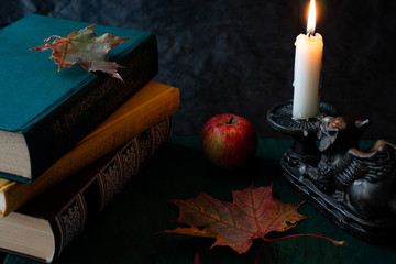 Naklejka na ściany i meble Autumn still life on a dark background with books, a vintage candlestick with a burning candle, leaves and a red apple.