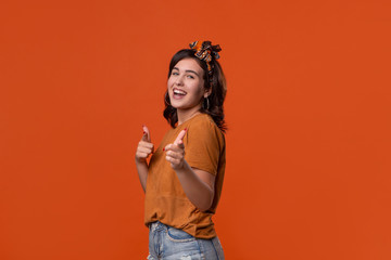 Obraz premium Smiling brunette woman in a t-shirt and beautiful headband pointing with her fingers at the camera isolated over orange background. Place for ad.