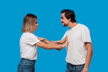 Fototapeta premium Couple of a young blond woman and brunet bearded man with mustaches in white t-shirts and blue jeans quarreling standing isolated over blue background. Concept of relationship crisis.