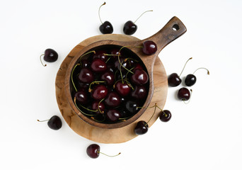 Top view: an image of a group of cherries in a wooden deep stewing pan, one berry on it, two cherries nearby and eight berries around on a wooden round stand (an isolated object) on a white background