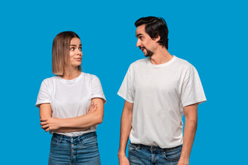 Fototapeta premium Happy couple of a young blond woman and brunet bearded man with mustaches in white t-shirts and blue jeans posing isolated over blue background. Concept of an ideal couple.
