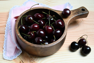 An image of a group of cherries in a wooden deep stewing pan, one berry on it, three cherries and a piece of pink fabric with a white and purple border nearby on a light colored wooden surface