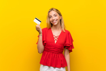 Blonde young woman over isolated yellow background holding a credit card