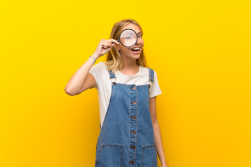 Blonde young woman over isolated yellow background holding a magnifying glass
