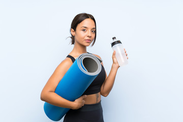 Fototapeta premium Young sport girl with mat and with a bottle of water over isolated blue background