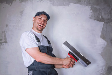 Portrait of a smiling worker in overalls and cap holding a putty knife in his hands against the...