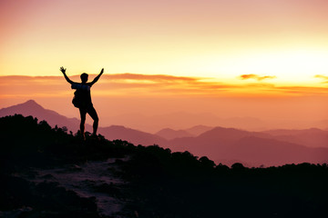 Hiker's silhouette stands on mountain top against sunset